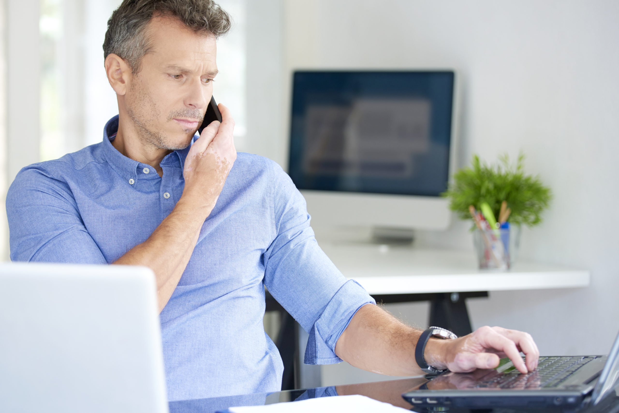 Portrait of middle aged businessman wearing shirt while sitting at the office and working. Professional man using his mobile phone and talking with somebody while working on laptops. Multitasking.